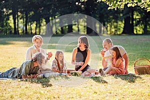 Big family sitting on the picnic blanket in city park during weekend Sunday sunny day. They are smiling, laughing and eating