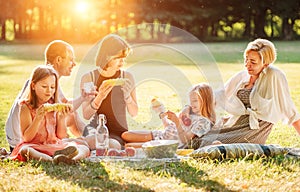 Big family sitting on the picnic blanket in city park during weekend Sunday sunny day. They are smiling, laughing and eating