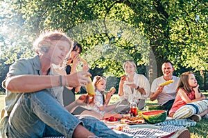 Big family sitting on the picnic blanket in city park during weekend Sunday sunny day. They are smiling, laughing and eating