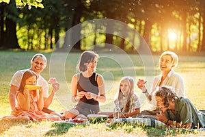 Big family sitting on the picnic blanket in city park during weekend Sunday sunny day. They are smiling, laughing and eating