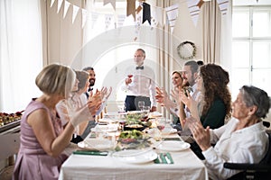 A big family sitting at a table on a indoor birthday party, a senior man giving a speech.