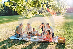 Big family under Linden tree on the picnic blanket on the in city park green grass. They are eating boiled corn, apples, peaches,
