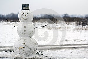 Big funny traditional primitive smiling snowman with bucket hat on white snowy field winter landscape, blurred black trees and blu