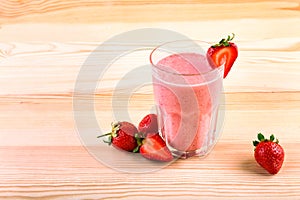 A big glass filled with an tasteful milkshake with a red strawberry on the top on a light brown wooden desk.