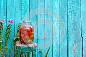 Big glass jar with pickled vegetables on chair on background of old wooden house. Canned tomatoes and cucumbers in