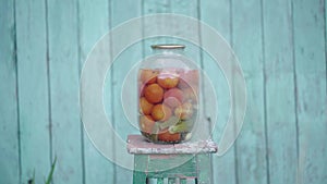 Big glass jar with pickled vegetables on chair on background of old wooden house. Canned tomatoes and cucumbers in