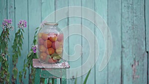 Big glass jar with pickled vegetables on chair on background of old wooden house. Canned tomatoes and cucumbers in