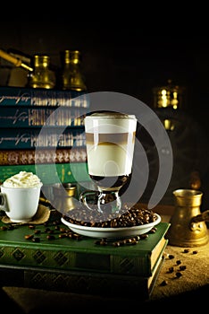 Big glass with latte stand in coffee beans beside turk. Atmosphere of library with old books around and grinder on dark background