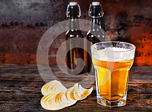 Big glass of light beer against two beer bottles near chips on d