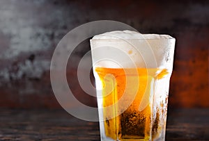 Big glass with a light beer and a head of foam on old dark desk