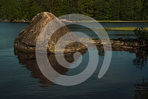 Big granite boulder in water of beautiful tranquil blue lake surrounded green forest in sunny summer day. Colorful summer Karelia.