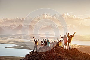 Grande gruppo di persone che hanno divertente nel successo posa con le braccia alzate sulla cima di una montagna contro il tramonto laghi e montagne.