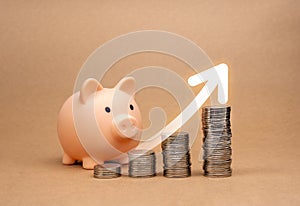 Big growing arrow rising on coin stacks as graph steps near cute pink piggy bank isolated on brown background. Saving money,