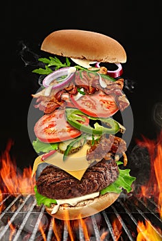 Big hamburger roasted on barbecue BBQ grill with bright flaming fire against black background. Beef cutlet, ham, cheese