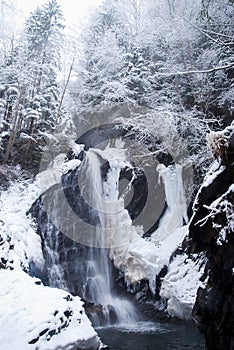 Big high Waterfall in the mountain winter forest with snow-covered trees and snowfall