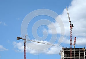 Big hoisting tower cranes and top section of modern construction building in a city over blue sky with clouds