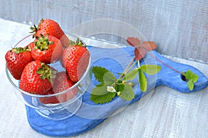 Big, homemade strawberry in transparent dish and blossoming flower on blue board