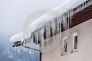 Big icicles and snow hanging over the rain gutter on a roof of a traditional wooden house in the mountains in winter.