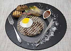 Big juicy grilled steak marbled beef with egg baked potatoes with barbecue sauce. Served on a stone plate with a fork and spoon on