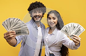 Big luck and win concept. Excited young indian couple holding dollar cash, posing with money on yellow background