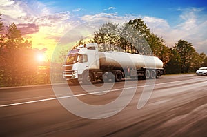 Big metal fuel tanker truck shipping fuel on the countryside road in motion with trees against sky with sunset