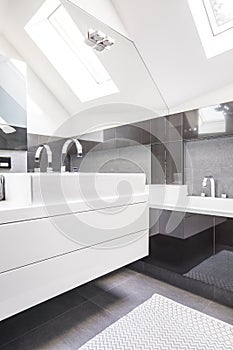 Big mirror by a modern, white washbasin cabinet in a fancy bathroom interior with reflective, black tiles and a roof window