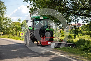 Big modern industrial tractor machine cutting green grass with mowing equipment along country roadside. Road lawn mower