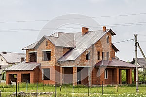 Big modern two-storied not finished new brick family cottage house with steep brown shingled roof, garages, high chimneys on blue