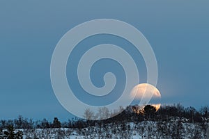 Big moon rising behind a mountain with some birch trees in winter landscape with snow, in Setesdal, Norway