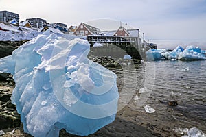 Big piece of blue ice laying among the stones on the shore with modern Inuit building on the hill at the fjord, Nuuk city,