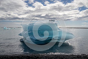 Big piece of blue ice over the black sand and sea water of Diamond beach, clouds on blue sky. Jokulsarlon - Iceland
