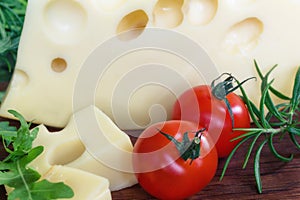 Big piece of cheese with holes lying with fresh twigs rosemary and cherry tomatoes on a wooden cutting board background.