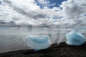 Big pieces of shinny and blue ice over the black sand of Diamond beach, clouds on blue sky. Jokulsarlon - Iceland