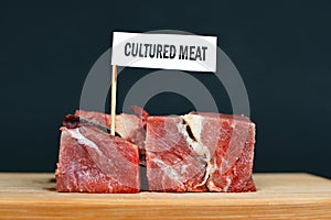 Big raw red meat chunks on wooden plate with label saying `cultured meat`