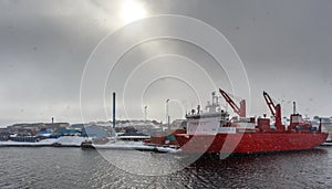 Big red cargo ship under the dull arctic sun and heavy snowfall in the port of Aasiaat, with village in the background, Greenland