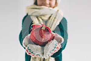 Big red Christmas ball in hands at the girl. The child is dressed in sweater, christmas hat and scarf studio shot.