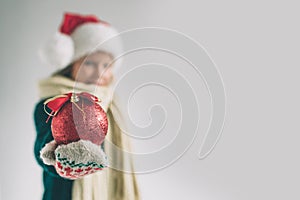 Big red Christmas ball in hands at the girl. The child is dressed in sweater, christmas hat and scarf studio shot