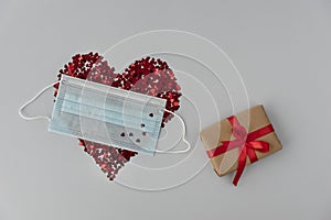 Big red heart made from little glittering confeti partly covered with medical mask little gift box is near, protective measures on photo