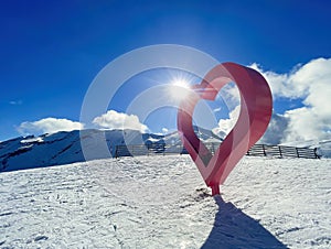 a big red metal heart in the middle of a ski slope, with snowy mountains and the sun behind, passion for skiing, winter sport,