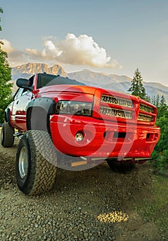 A big red monster truck with dirt tires standing on an off road against mountains and dramatic clouds