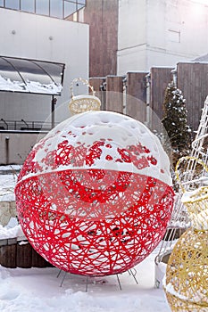 A big red snow-covered Christmas ball made of metal wire on the street of a big city on a sunny winter day.