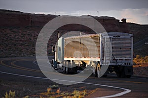 Big rig semi truck with bulk covered semi trailer going on winding road with red rocks of Arizona landscape in twilight