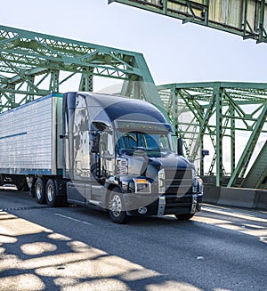 Big rig black stylish diesel semi truck transporting cargo in refrigerator semi trailer running on the arched truss Interstate