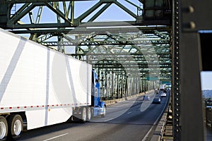 Big rig blue classic semi truck transporting goods in refrigerated semi trailer driving on the metal truss interstate highway