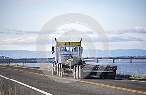 Big rig blue day cab semi truck transporting empty step down semi trailer for oversized loads running on the narrow local road