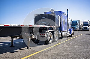Big rig blue semi truck with empty flat bed semi trailer standing on truck stop parking lot opposite another semi trucks standing