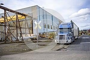 Big rig blue semi truck with grille guard standing with open semi trailer on the road shoulder in industrial area waiting for next