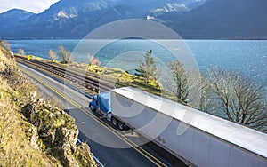 Big rig blue semi truck with refrigerated semi trailer transporting goods running on the road along railroad in Columbia Gorge