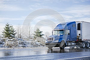 Big rig blue semi truck tractor transporting commercial cargo in refrigerator semi trailer going on the wet road with melting snow