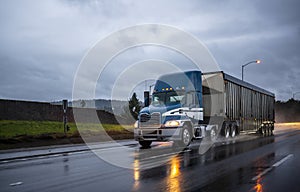 Big rig bonnet blue semi truck transporting cargo in covered bulk semi trailer running on the wet glossy road with raining weather